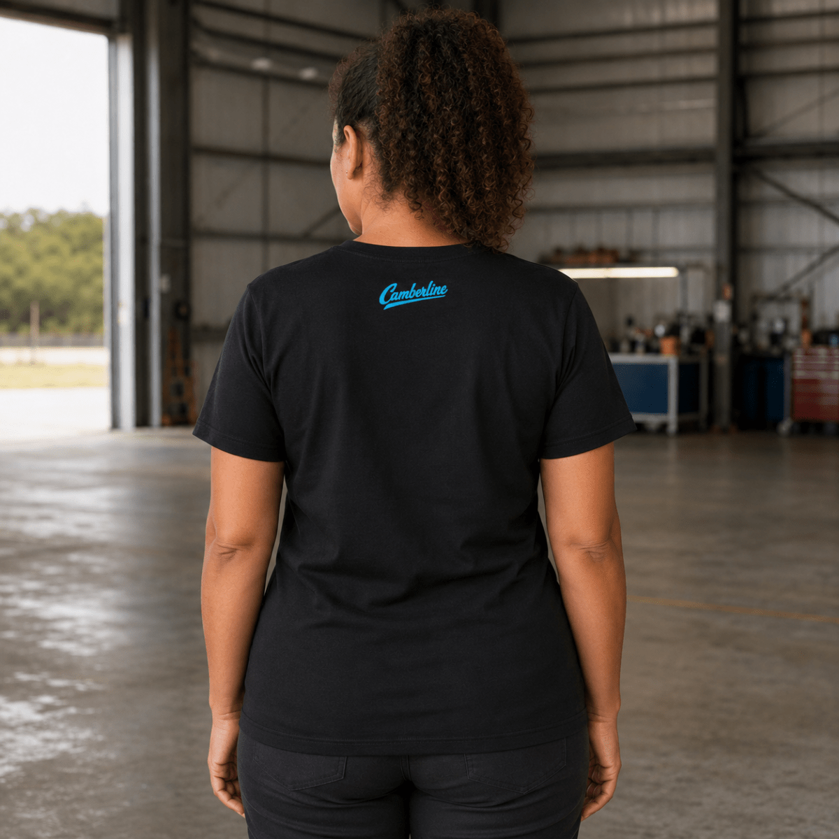 A woman viewed from behind wearing a black short-sleeve t-shirt with a small blue 'Camberline' logo on the back, standing inside a large industrial garage or workshop.