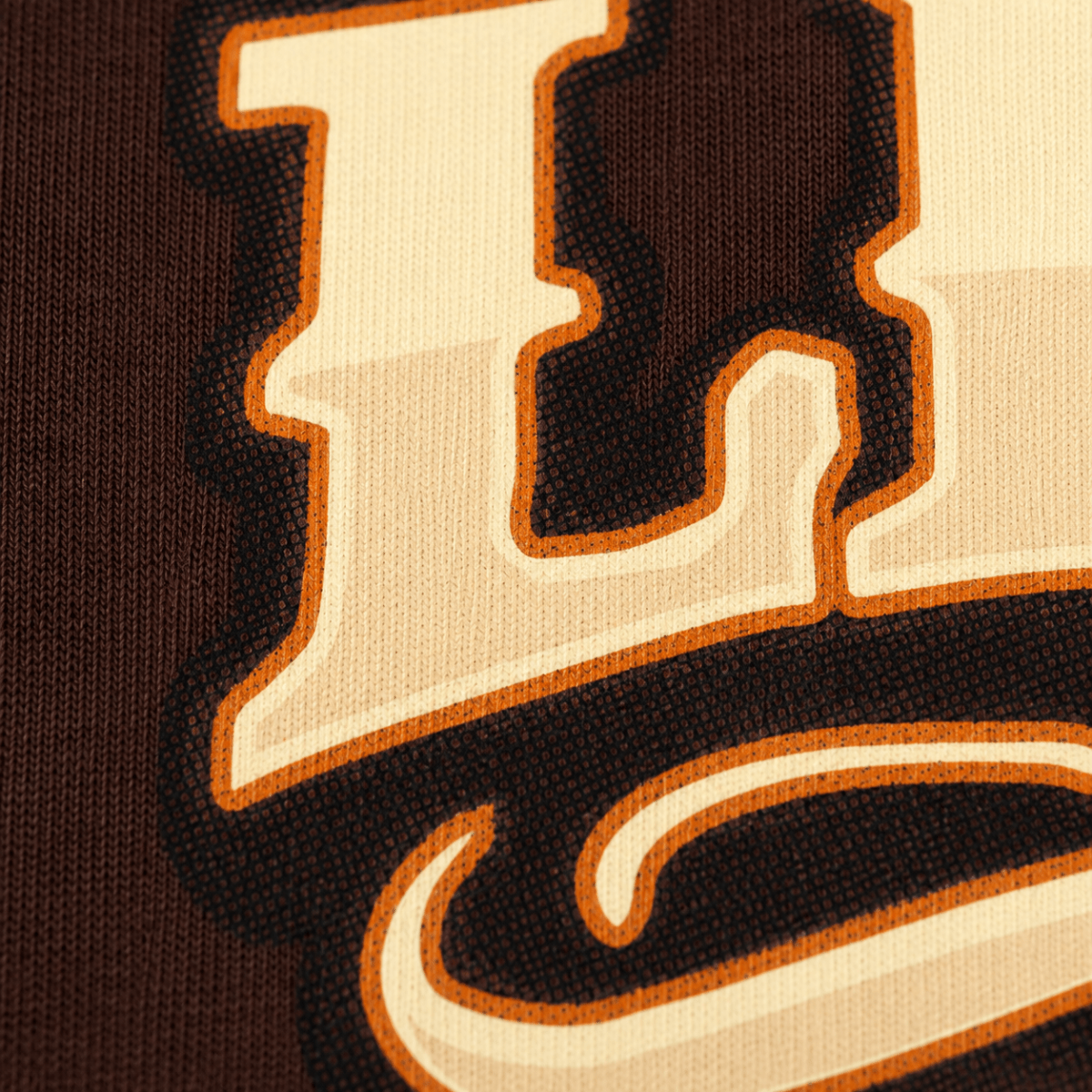 Close-up of a knit fabric logo patch featuring a large cream letter with a dark brown fill and orange outline on a dark brown background.
