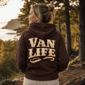 A woman stands outdoors on a wooded path at sunset, shown from the back wearing a brown hoodie with large cream lettering that reads “VAN LIFE.”.