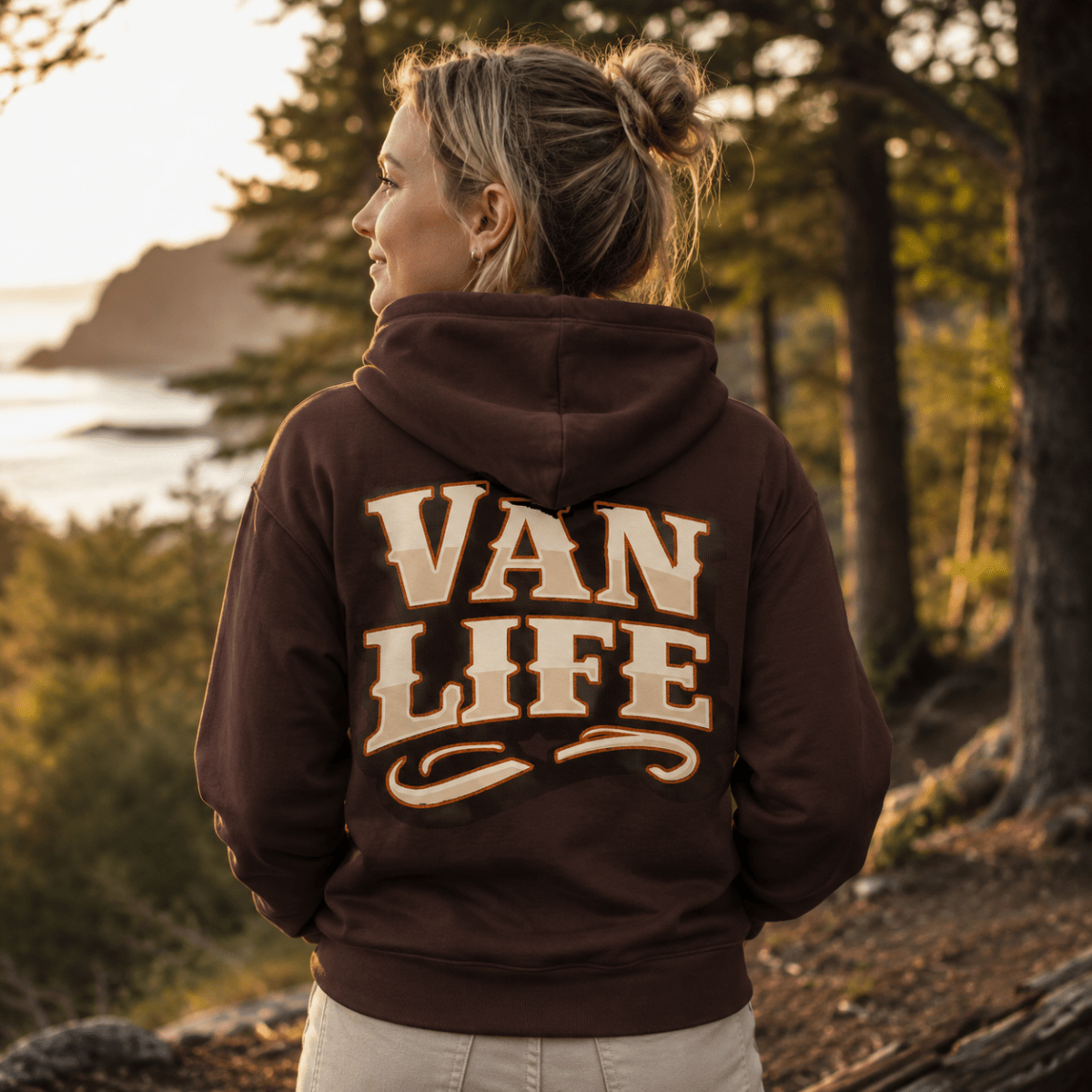 A woman stands outdoors on a wooded path at sunset, shown from the back wearing a brown hoodie with large cream lettering that reads “VAN LIFE.”.