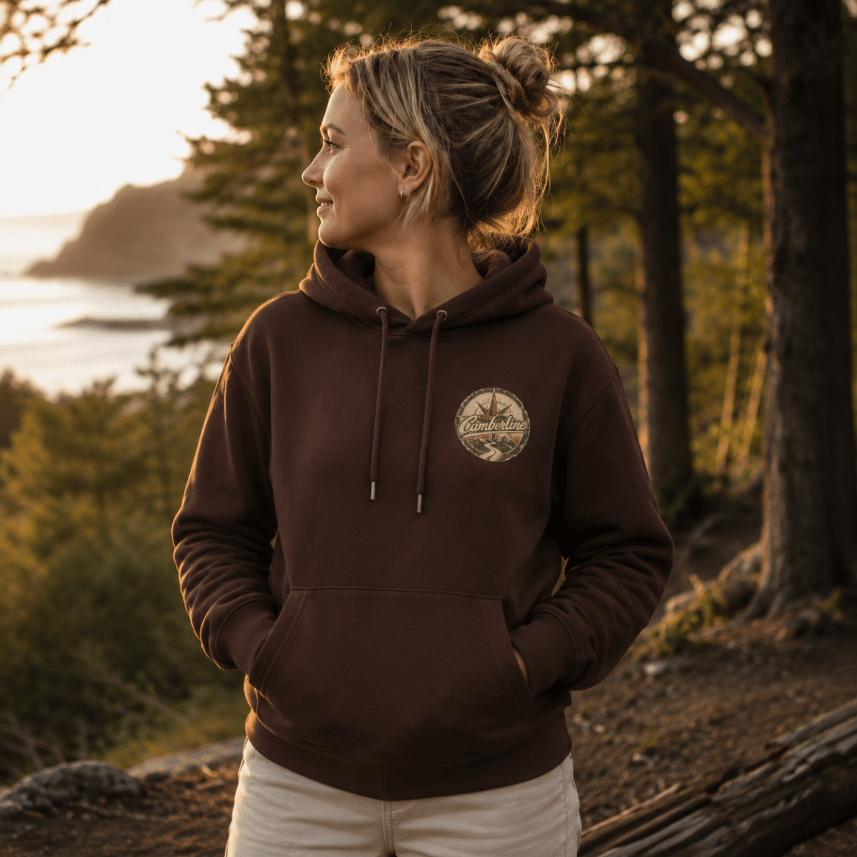 A woman wearing a dark brown pullover hoodie with a circular chest graphic, standing outdoors in a forest with a coastal view in the background.