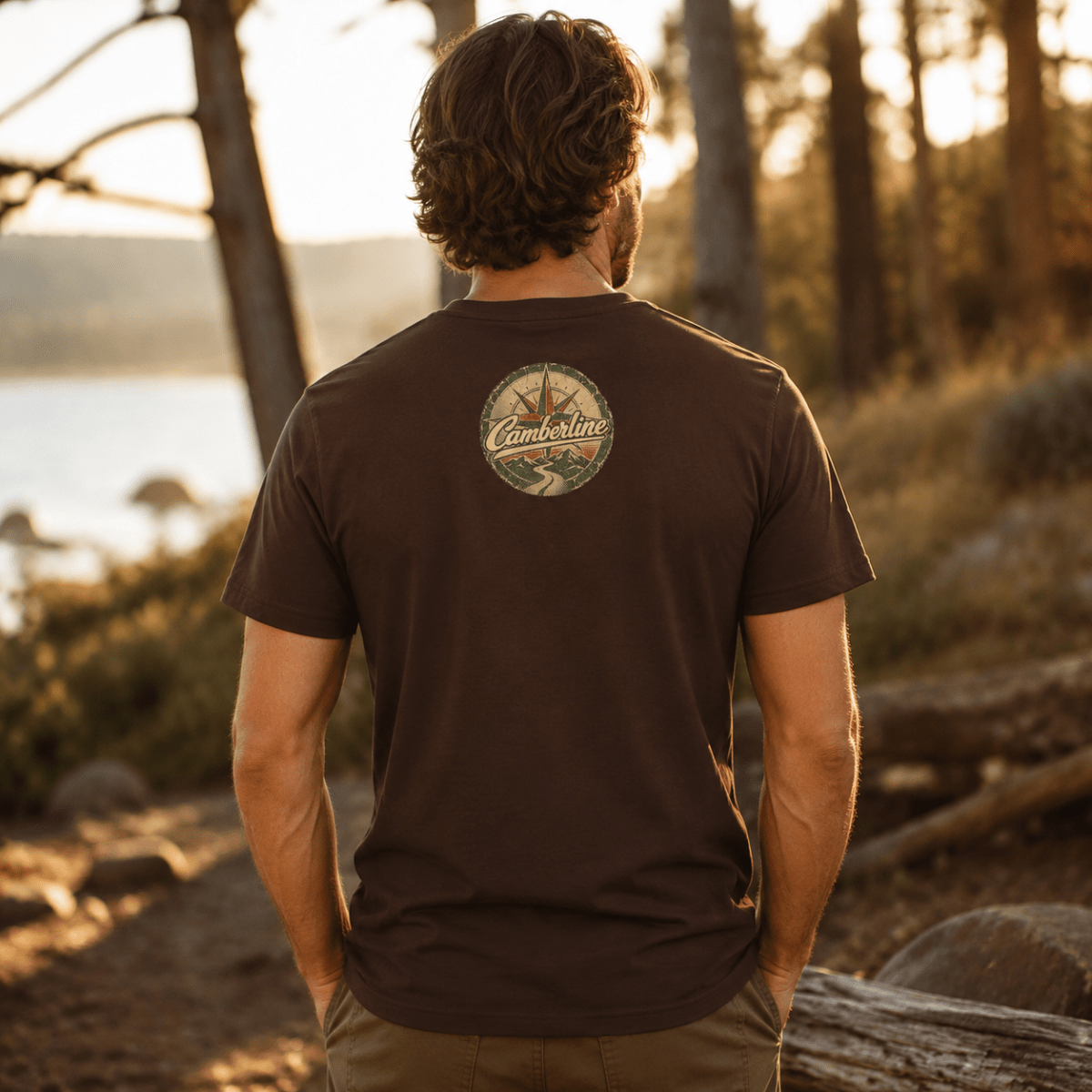 A man viewed from behind wearing a dark brown t-shirt with a circular 'Camberline' logo printed on the upper back, set against a warm, golden outdoor forest and lake background.