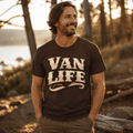 A man wearing a dark brown t-shirt with 'VAN LIFE' printed in a cream-colored retro font, standing outdoors in a wooded setting during golden hour.